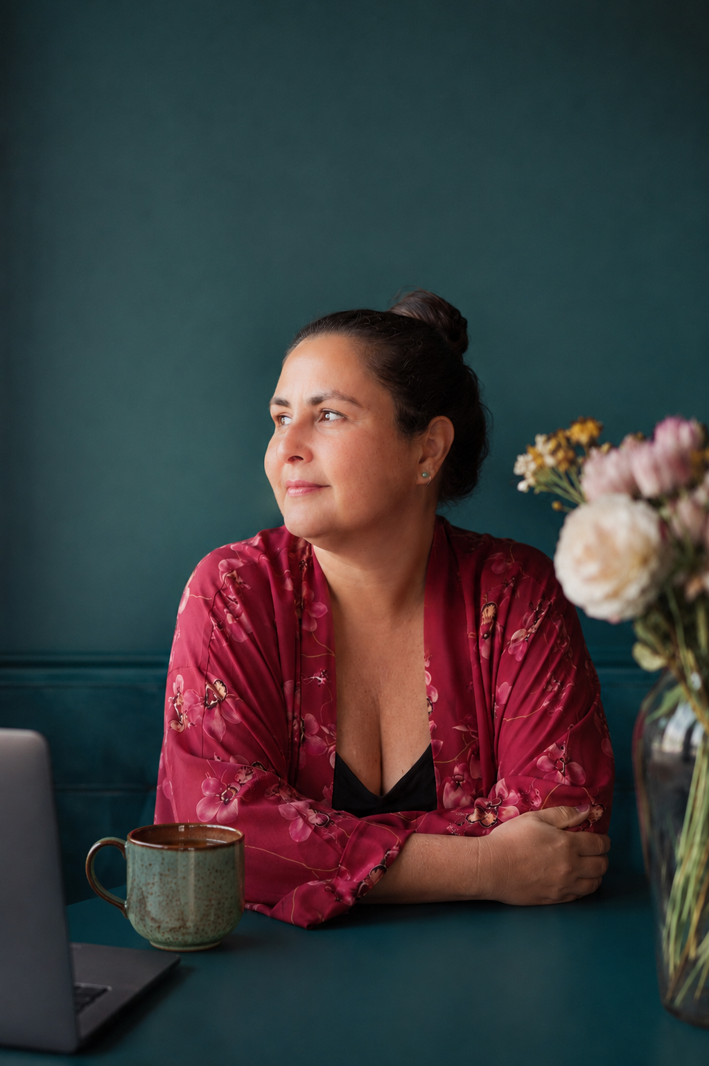 Cris apoiada na mesa, em luz natural, ao lado de um laptop e um vaso de flores